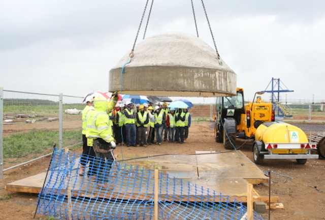 Foreground - a concrete dome is lifted by an excavator; background, twenty students look on under umbrellas